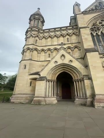 The video shows the exterior of a large, light-colored stone building that appears to be a cathedral or church. The building has intricate architectural details, including arched doorways, a rose window, columns, and a spire. The sky is overcast. A woman in a long dark dress and black jacket walks into the frame and stops in the paved courtyard in front of the building. She faces the camera, puts her hands together in front of her chest, and smiles.