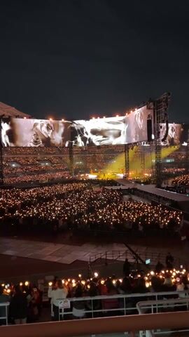 The video shows a crowded concert at night. People in the audience are holding lights, creating a sea of illumination. The stage has large screens displaying visuals. There is music playing and people seem to be enjoying the show.