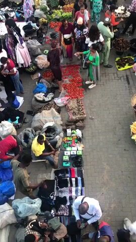 The video shows a crowded outdoor market. Numerous people are walking around and browsing stalls. There are many different types of goods for sale, including produce such as tomatoes and corn, as well as clothing and other items. Some people are interacting with vendors, and others are carrying bags or baskets. The ground appears to be paved with bricks. The lighting suggests it is daytime, possibly late afternoon due to the shadows. The overall atmosphere is busy and active.