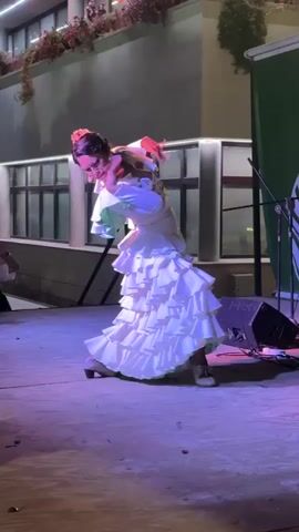 A woman in a white ruffled dress dances on a stage. She has her hair up and is wearing a headdress. She is facing away from the camera, with her left arm raised and her right hand near her chest. The stage is lit with purple and pink lights. Behind her, a building with windows and plants can be seen. To the right of the stage, there is a speaker and a green banner. A person with a camera is visible in the lower left corner of the frame, looking at the dancer. The woman's dress has multiple layers of ruffles, and it flares out as she moves. Her shoes appear to be heeled. The overall scene depicts a performance, likely a flamenco dance, taking place outdoors at night.