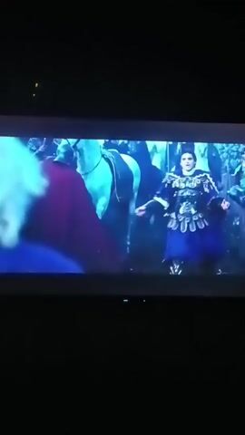 The video shows a scene from a film displayed on a television. In the foreground, a person with blonde hair is seen from behind, facing away from the camera. To their right, a man in a blue and gold patterned tunic and armor stands with his arms outstretched. Behind them, several horses and people are visible in a dimly lit outdoor setting, possibly a medieval or fantasy environment. The scene shifts to a close-up of a man in armor, looking down with a pained expression. Then, the view returns to the initial scene, focusing on the interaction between the blonde-haired person and the man in the ornate tunic.