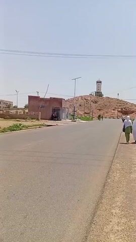 A wide shot of a paved road under a clear sky. On the left side of the road, a person in a blue and green outfit is sweeping with a broom and gathering debris into a white bag. Behind them are a few buildings and a small tree. Further down the road, two people wearing white robes walk along the sidewalk. In the background, a lighthouse stands atop a rocky hill, with a few other structures and people visible around its base. The environment appears dry and sunny, with some sparse vegetation along the roadside.