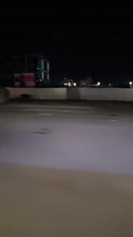 The video is taken from a moving vehicle at night. The camera is focused on the road ahead. On the right side of the road, there is a long, low wall or barrier. Beyond the barrier, there are blurry structures including what appear to be unfinished buildings and a trailer. Street lights illuminate the area in the distance. A post with yellow and black stripes passes by.  The road is paved and appears relatively smooth. The overall impression is of a desolate or industrial area.