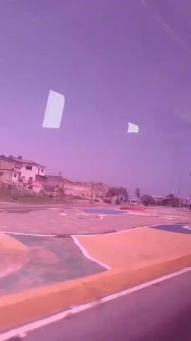 The video, filmed from inside a vehicle, shows a view through a window of a street and buildings. The sky is a purplish hue, with two white reflections on the window. In the foreground, a yellow curb is visible, separated from the street by a white line. Behind the curb is a multi-colored paved area with sections of blue, pink, and white. Further back, there are low-rise buildings with some vegetation. A white bus is parked in the distance. The scene appears to be outdoors in daylight.