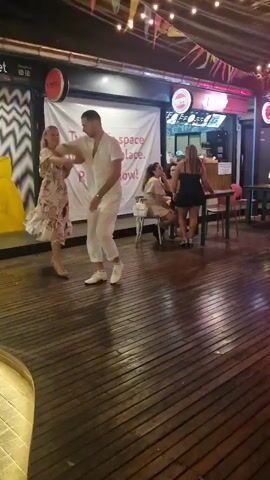 A couple dances on a wooden floor in front of a marketplace. The woman wears a floral print dress and the man wears a white outfit. In the background, other people are seated at tables. The marketplace has posters on the wall, one featuring a stylized image of Mona Lisa eating noodles. String lights hang overhead.