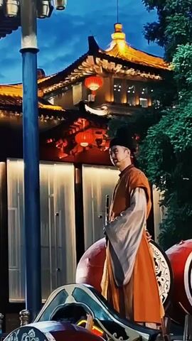 An eye-level shot captures a man standing in a traditional Chinese outfit, against a backdrop of Chinese architecture and a darkening blue sky. The man is dressed in an orange robe over a white shirt, with long, flowing sleeves. He wears a black hat and holds a long, thin object, possibly a musical instrument, in his hands. He is standing on a decorated platform with red and black accents. Behind him, a traditional Chinese building is visible, adorned with red lanterns and glowing lights. The building has a classic curved roof design. The sky transitions from a lighter blue to a darker shade, indicating either dusk or dawn. To the left of the man is a tall, blue lamppost with multiple lights, and to the right is a dense green tree. The overall scene evokes a sense of cultural performance or festival in a traditional Chinese setting.