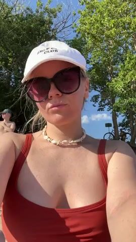 A young Caucasian woman with blonde hair is shown in a close-up selfie. She is wearing a white baseball cap with text that appears to read "SELF-LOVE CLUB". She has on large, dark sunglasses and a seashell necklace. Her top is a reddish-orange color, likely a swimsuit. She is looking directly at the camera with a neutral expression. In the background, out of focus, is greenery from trees, a blue sky with some clouds, and other people and objects, including what appears to be a bicycle. The lighting suggests it is daytime and sunny.