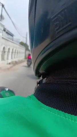 The video shows a first-person perspective from someone riding on the back of a motorcycle, wearing a green jacket with a logo that appears to be a stylized bird with wings spread. The rider's helmet is partially visible in the foreground. The motorcycle is traveling on a road with other vehicles, predominantly other motorcycles. On the left side of the road, there is a white wall with arched details, followed by some trees and utility poles with many wires. On the right side of the road, traffic includes several motorcycles, one of which has a driver wearing a red helmet and a backpack, and further ahead, a large truck with a shipping container. The sky is overcast. The video captures a slice-of-life scene of traffic on a street, likely in a Southeast Asian country given the presence of motorcycle-heavy traffic and the Gojek-like logo.