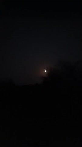 A video shows a nighttime outdoor scene. The camera pans slightly to the left, revealing a dark silhouette of trees and vegetation against a dark sky. A bright, circular light source, likely the moon, is visible near the center of the frame, appearing somewhat hazy due to atmospheric conditions or possibly light pollution. The darkness dominates the majority of the video, with the moon being the primary point of illumination.