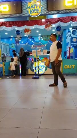 A man in a white t-shirt and dark pants walks away from the camera in a brightly lit indoor space. Behind him, a "Happy Land" play area is visible with blue walls decorated with planets and a cartoon spaceship. Several children and an adult are standing at a counter in the play area, seemingly interacting with staff. A sign with the word "OUT" is visible to the right of the play area. The floor is tiled with large beige squares.