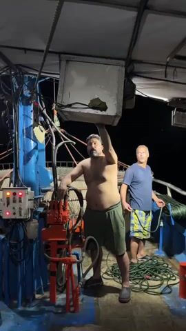 Two men are on a boat at night. The man in the foreground is shirtless and wearing shorts, holding a white rectangular object over his head with his right arm. He is standing next to a red and orange piece of machinery. The man in the background is wearing a t-shirt and shorts, standing near a coiled green rope. The scene appears to be on a fishing vessel, with various cables and equipment visible.