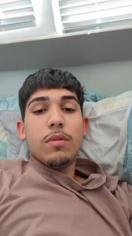 A young man with dark hair, a mustache, and a goatee, wearing a brown hooded shirt, lies on a bed with patterned pillows. He gestures with his right hand and then smiles while looking directly into the camera.