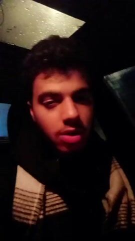 A man with dark hair and a light complexion covers his mouth with his hand in a dark environment, possibly inside a car, as a seat is visible in the background. He is wearing a dark top and a scarf with a striped pattern. The lighting is dim, casting shadows across his face.