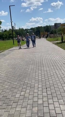 A paved walkway stretches towards a crowd of people in the distance on a sunny day. Two women and a boy walk towards the camera on the left side of the walkway. In the background, people are gathered in a park with trees and buildings. A yellow swing set is visible on the right side of the frame.