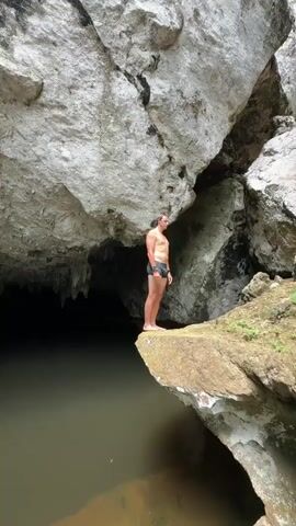 In this video, a man is captured performing a backflip from a rocky ledge into a dark cave pool. The setting appears to be a natural cave with a large rock overhang and stalactites hanging from the ceiling. The water is murky, and the light is dim, suggesting an indoor or shaded environment. The man is wearing dark swimming shorts and is shirtless. He is seen standing on the edge of the rock, crouching, and then mid-air in the process of flipping. The video concludes with him splashing into the water, creating a significant disturbance on the surface. The overall atmosphere is adventurous and daring, highlighting the thrill of cliff jumping in a natural, somewhat mysterious location. The natural lighting conditions suggest it could be daytime, but the cave environment makes it difficult to determine the exact time of day. There is no visible text in the video.