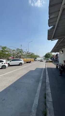A vertical video captures an outdoor scene on a bright, sunny day. The camera is positioned at a low angle, looking upwards towards the sky and down a paved road. On the left side of the road, several white cars are parked or in motion. A white SUV is prominent in the foreground. Further down the road, a yellow truck is visible. On the right side of the road, there is a sidewalk with a shaded overhang from a building, supported by grey pillars. A person is sitting on a dark bench on the sidewalk, partially obscured by shadow. The background features a clear blue sky with a few scattered clouds, green trees, and utility poles. The road surface appears to be asphalt or concrete. The overall impression is a street or parking area with some traffic and pedestrian access.