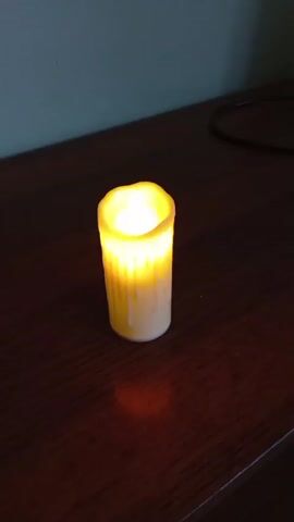 The video displays a close-up shot of a single, white LED candle with a flickering yellow light. The candle is positioned on a dark, polished wooden surface, possibly a table or shelf. A faint orange glow emanates from the candle and reflects on the surface beneath it. In the background, a dimly lit, pale blue wall is visible, with a thin wire or cable extending from the upper left corner downwards towards the candle. The overall lighting is dim, emphasizing the warm glow of the candle. The camera remains stationary throughout the clip.