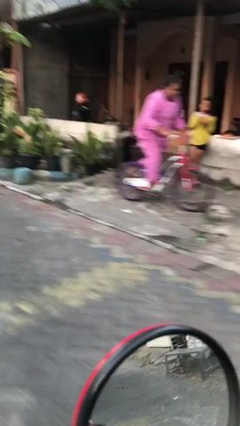 A young woman in a pink outfit rides a bicycle. The bicycle has a red frame, white handlebars, and purple wheels. The woman is in motion, pedaling the bicycle. The background shows a residential area with a house. The ground is paved with bricks.