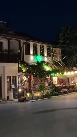 The video shows a street scene at night. A building with white walls and a tiled roof is decorated with numerous strings of small lights, some green and some white. There are also larger, yellow lights illuminating the facade and what appears to be an outdoor seating area. Plants and flowers are visible in planters along the front of the building. A sign with red lettering is hanging above the entrance. Several tables and chairs are set up outside, suggesting a restaurant or cafe. Further down the street, other buildings are visible, also illuminated by lights. A street lamp is visible on the right side of the road. There are a few cars parked or driving on the street, and a traffic sign is visible. The sky is dark, indicating it is nighttime.