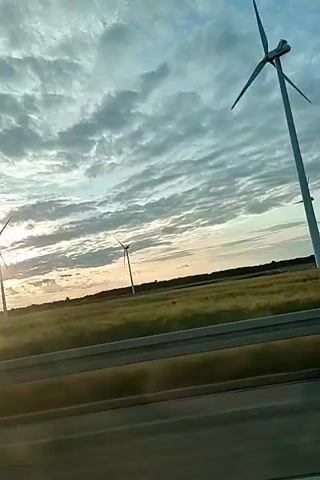 The video shows a view from a moving vehicle looking out at a landscape with several wind turbines. The sky is partly cloudy with the sun visible, suggesting either sunrise or sunset. The wind turbines are tall structures with rotating blades, characteristic of renewable energy generation. A highway or road is visible in the foreground, with a metal barrier separating it from the field. The field appears to be covered with dry grass or crops. The overall impression is of a journey through a rural area during a time of day with dramatic lighting.