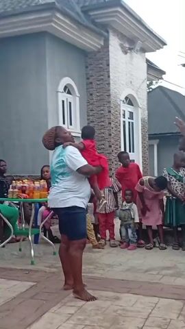 The video captures an outdoor gathering of adults and numerous children on a paved courtyard in front of a modern, light-colored house with stone cladding. A large group of children, dressed in various casual outfits, are actively engaged with an adult, raising their hands and appearing to participate in a game or activity.

In the initial frames, an adult wearing a light blue and green tie-dye shirt and dark shorts, with a patterned head covering, holds a baby while facing a semicircle of children. Another adult in a white top and dark pants is seated at a table laden with bottled drinks and cups. As the camera pans, more adults are seen seated around the table, conversing and observing the scene. One person wears a white sleeveless top, another a black t-shirt, and a third a pink top. A blue plastic chair and a white cooler are visible near the table, along with a white bag with "FOR SHOPPING" printed on it.

Later in the video, a different adult, dressed in a blue short-sleeved top or dress, stands facing a similar group of children, who again have their hands raised, indicating continued engagement in the activity. The children appear happy and animated, while some adults are seen smiling and laughing. The background features the house with arched and rectangular windows, bushes, and a water tank on a metal stand. The setting suggests a communal event or party during daytime, likely in a warm climate, consistent with the provided context of Onicha, Nigeria.