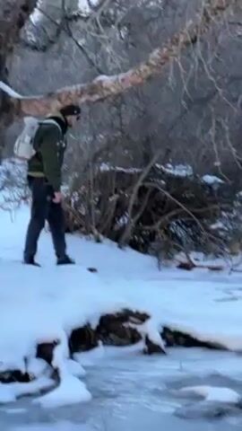 A man in a green jacket, dark pants, and a gray backpack stands near a partially frozen stream in a snowy environment. He is bent over, looking at something in the water. The scene is wintery, with bare trees and snow-covered ground. Someone off-camera speaks in a language other than English.