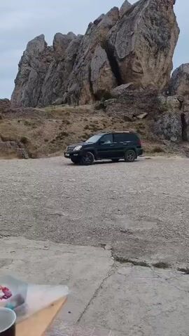 A black SUV is parked on a gravel lot in front of a large rock formation under a cloudy sky. The SUV is angled slightly to the left. The ground is a mixture of concrete and gravel. In the foreground, a corner of a wooden table and some cups are visible. The rock formation is jagged and appears to be made of light-colored stone. The overall scene suggests an outdoor location, possibly a tourist spot or a scenic viewpoint.