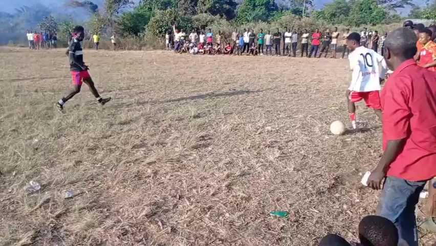 A group of people are gathered in a dry, grassy field, watching a soccer game. Two young men are actively playing, one in a black and red uniform and the other in a white and red uniform. A soccer ball is visible on the ground between them. In the background, more people are lined up, observing the game, and trees and mountains form the backdrop. There is some smoke in the distance to the left. The overall scene suggests an informal outdoor sporting event in a rural or underdeveloped setting.