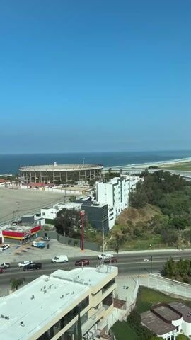 The video shows an exterior view of a city from a high vantage point, likely a hotel window. The scene is dominated by a bright blue sky and the ocean stretching to the horizon. Below, a stadium is visible along the coast, with a large parking lot in front.  Several buildings, including a convenience store and apartment complexes, are scattered throughout the area. Cars move along the roads, and palm trees add to the coastal atmosphere.  The edge of the window frame becomes visible, indicating the perspective is from inside a building.