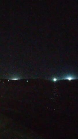 The video depicts a night scene by the water. A dark shoreline is visible in the foreground, with faint lights scattered along the horizon in the distance. The water reflects the lights, creating a shimmering effect. The overall atmosphere is dark and quiet, with the primary focus on the contrast between the lights and the darkness.