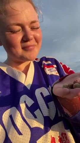 A young woman with her hair pulled up in a bun and wearing a blue and white hockey jersey with Russian text and symbols is shown in a close-up shot. She has a nose ring and earrings. Her eyes are closed and her mouth is slightly open, suggesting she might be singing, speaking, or experiencing a strong emotion. The jersey has "РОССИЯ" prominently displayed in large white lettering. On the right chest of the jersey, there is a red, white, and blue crest with text that includes "СССР" and "Российские Звезды".

The background shows a slightly blurred landscape with green trees and a cloudy sky, illuminated by the warm glow of what appears to be sunset or sunrise. The lighting suggests it is either early morning or late evening. The setting appears to be outdoors in a natural environment, possibly a hill or meadow overlooking a wooded area, consistent with a rural location like Olkhovka, Russia. The woman's expression conveys a sense of peace or reflection.