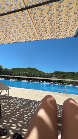 The video shows a first-person perspective of someone relaxing by a swimming pool on a sunny day. In the foreground, the person's legs are visible, positioned on a lounge chair under a patterned sunshade. The sunshade casts a dappled shadow on the ground. Beyond the lounge chair, a large, bright blue swimming pool is in view, with steps leading into it. Several lounge chairs are arranged around the pool, some occupied by people sunbathing or sitting. The background features a lush green hillside dotted with trees and buildings. The sky is a clear, vibrant blue with a few wispy clouds. The overall scene depicts a leisurely and pleasant atmosphere at a resort or a similar recreational facility.