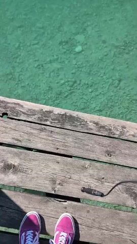 The video shows a pair of pink sneakers standing on a wooden dock next to clear turquoise water. The water is shallow, with rocks visible on the bottom. A black wire is seen partially submerged in the water, moving with the current.