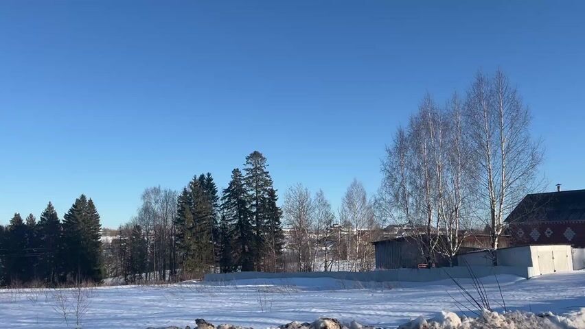 The camera slowly pans across a snowy winter landscape under a clear blue sky.  The landscape features snow-covered fields and rolling hills. A line of bare and evergreen trees borders one side of the field.  Wooden telephone poles with wires stretch across the landscape. Buildings, including small sheds and houses, are visible in the background. The scene appears peaceful and rural.