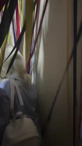 A person with blonde hair wearing a light-colored hoodie and a backpack walks through a narrow passage decorated with colorful streamers. The passage is dimly lit, and the streamers hang down from above, creating a visually stimulating environment. The person appears to be moving away from the camera and deeper into the passage.