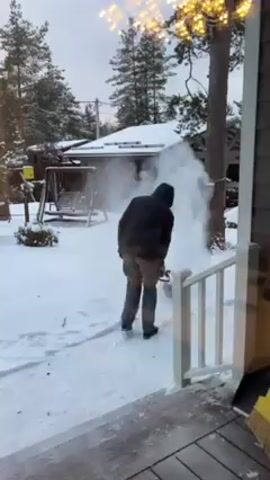 A man in a dark coat and gray pants shovels snow in a snow-covered yard. The camera is positioned on a porch, looking out over the snowy yard. A white railing of the porch is visible on the right of the frame. A swing set, several trees, and a building are in the background, all covered in snow. A string of lights hangs above the camera. The man is actively shoveling snow, and plumes of snow are being dispersed by the shovel.
