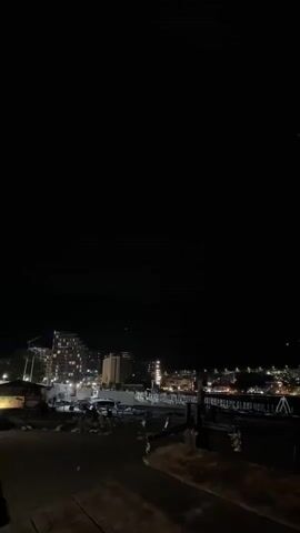 This video presents a nighttime view of a coastal area with illuminated buildings and a dark sky. The foreground shows a sandy or gravelly surface, suggesting a beach or waterfront promenade. Several wooden structures and what appear to be posts or pilings are visible.

In the middle ground, there is a collection of modern buildings, some of which are hotels or apartment complexes with many lit windows. Construction cranes can be seen behind some of these structures, indicating ongoing development. A few cars are parked or driving in this area, their headlights contributing to the illumination. A pier or boardwalk extends into the water, also lit with strings of lights.

The background features a dark silhouette of mountains against the night sky, and further along the coast, more lights from a town or city can be seen. The overall scene evokes a sense of a lively, developed coastal town at night. The time of day is clearly night, indicated by the darkness and artificial lighting. No specific weather conditions are apparent, though the sky appears clear enough to see distant lights.

There is no visible text in the video. The mood is peaceful and somewhat serene, with the lights creating a warm glow against the darkness.