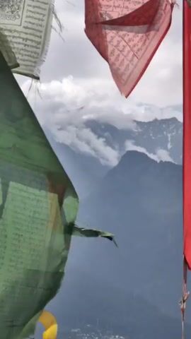 The video captures a serene mountain landscape with prayer flags in the foreground. The flags, in colors like green, red, and white, are unfurling and fluttering gently in the wind. Behind the flags, a majestic mountain range is visible. The mountains are layered and shrouded in a soft mist or clouds, with some snow-capped peaks peeking through. The sky is overcast with a mix of gray clouds. The overall impression is one of tranquility and natural beauty, with a spiritual undertone suggested by the presence of the prayer flags.
