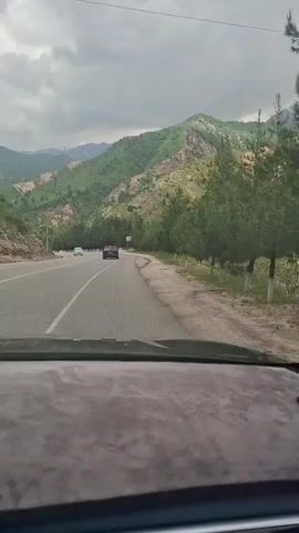 The video shows a car driving on a highway in a mountainous area on an overcast day. The road is paved with a white line down the center. There are several cars on the road, and green trees line the side of the highway. The mountains in the background are green with some bare patches. The interior of the car is visible from the dashboard up. The audio plays pop music.