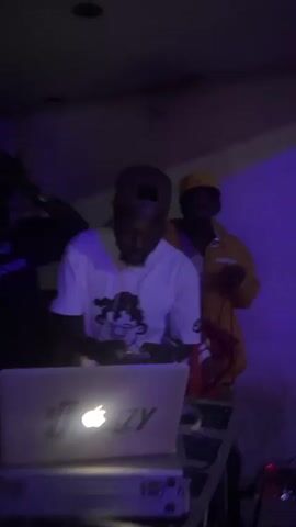 The video shows a dimly lit indoor scene, possibly a party or club. In the foreground, a person wearing a white t-shirt with a cartoon graphic and a baseball cap is standing behind a DJ setup, which includes a laptop with an Apple logo and DJ controllers. The person appears to be operating the DJ equipment and holding a phone. In the background, another person wearing a yellow hoodie and a cap is holding a microphone and singing or speaking into it, illuminated by a red light from the microphone. Another individual, wearing a dark t-shirt, is partially visible in the background, also holding a microphone and appearing to sing. The environment is dark with some purple and pink colored lights.