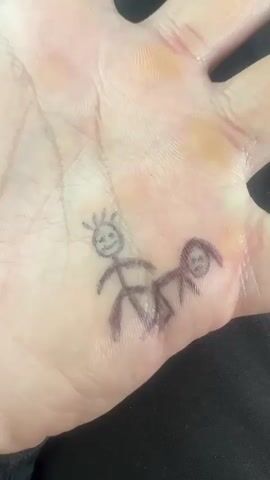 A close-up video shows the palm of a human hand. Drawn on the center of the palm are two simple stick figures in black ink. The left figure is a full body stick person with spiky hair and a smiling face. To its right, and slightly below, is another figure depicted as a head in profile with some hair, facing away from the first stick figure. The hand itself appears light-skinned and shows some subtle reddish discoloration or calluses, particularly near the base of the fingers and thumb. The background is a dark, blurry fabric, possibly clothing. The video is very short, showing the hand and drawings with a slight, almost imperceptible wobble. There is no visible text.
