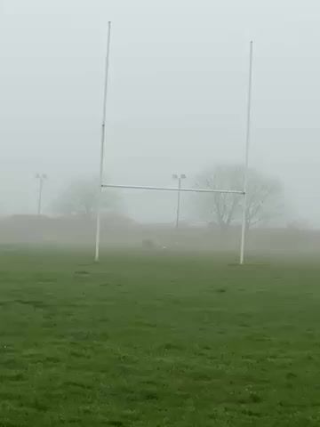 A person is kicking a rugby ball on a foggy day on a green field. Rugby goal posts are in the center of the frame, and the person is behind them, further in the distance. There are trees and light posts in the background, barely visible through the fog.
