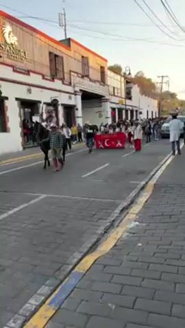 The video shows a parade moving down a cobblestone street in a town. On the left side of the street is a two-story building, white with red trim. The first person is riding a horse. Next to the horse, a man walks wearing a plaid shirt. The parade is led by people carrying a large banner with Turkish symbols on it. Behind them, people are walking and playing musical instruments. People are watching the parade from the sidewalks. At the end of the street is a white SUV. The sky is blue.