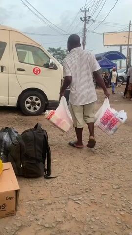 The video begins with a shot of a white van with a red logo on its side, parked on a dirt road. In the foreground, a black backpack and a brown cardboard box are visible. A man in a striped shirt and shorts is walking away from the camera, carrying two plastic shopping bags. Behind him, another man in a green shirt and shorts walks towards the van. In the background, there are more vehicles, people, market stalls, and utility poles. The sky is overcast. The camera pans slightly to the right, following the man with the shopping bags as he moves further away. Another man in a green shirt is seen near the van, and a woman is walking in the distance. The scene appears to be a busy outdoor marketplace or transportation hub.