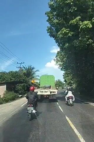 The video shows a view from a vehicle traveling on a paved road. In front of the vehicle, there is a white truck with a large green object secured on its flatbed. Two motorcycles are also on the road, one directly in front of the truck and the other to the right of the truck. Both motorcycles have riders. The road is lined with trees and some buildings on the left side. The sky is blue with some white clouds. The overall environment appears to be a rural or semi-rural area with clear weather.