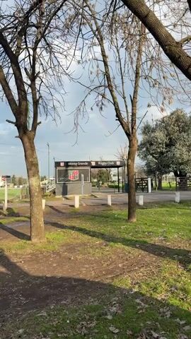 The video shows a view of a rugby club entrance with trees in the foreground. A small building with a sign that reads "Monte Grande Rugby Club" is visible. A faint rainbow is present in the sky. The ground is covered with grass and dirt. The scene appears to be outdoors during the daytime.