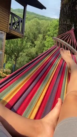 The video shows a first-person perspective of someone relaxing in a colorful striped hammock outdoors. Their feet are visible, resting on the hammock. In the background, there is a wooden structure resembling a treehouse or cabin, lush green trees, and a hillside. The lighting suggests it is daytime, with sunlight casting shadows. The camera is stationary, capturing a peaceful, natural scene.