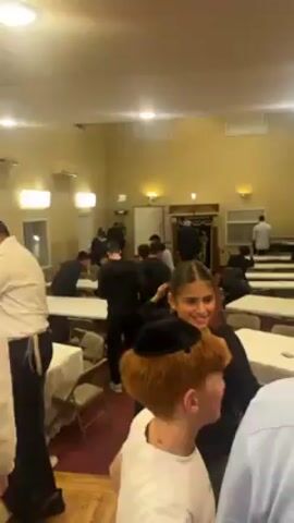 The video shows the interior of a large room with light yellow walls and a red patterned rug. There are numerous people, mostly young men and boys, some wearing yarmulkes. Many tables covered with white cloths are arranged in the room, and some people are seated at them. Some people are standing and talking, and one man is shown eating from a small white dish. There are windows with blinds on some walls, and a built-in bookshelf with a dark wood finish is visible in one corner. The room is well-lit by overhead fluorescent lights and sconces on the walls. The camera moves slightly, providing a wider view of the room and the people within it.