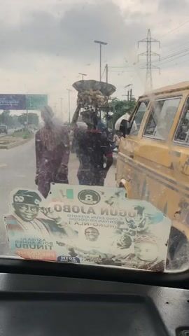 The video shows a view from inside a vehicle looking through the windshield. The windshield is covered in smudges and dirt. A sticker with images of political figures is affixed to the lower part of the windshield. In the background, there are people walking on a street, a yellow van parked on the right side, streetlights, and utility poles. One person in the background is carrying an object on their head. The sky appears overcast.