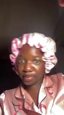A young woman with dark skin wearing a pink satin bonnet and matching pajamas is looking directly at the camera. Initially, she is speaking with her mouth open and appears agitated. Subsequently, she purses her lips, puckering them. The background is dark and indistinct.